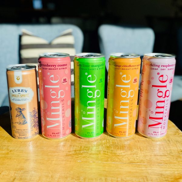 Five colorful beverage cans, including one Lyre's Amalfi Spritz and four Mingle mocktails, are lined up on a wooden surface with a soft-focus background.