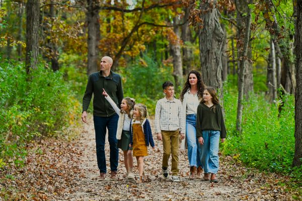 A family of six walks together on a leaf-covered path in a forest during autumn, surrounded by green and brown trees.