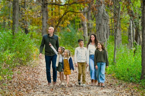 A family of six walks together on a leaf-covered path in a forest during autumn, surrounded by green and brown trees.