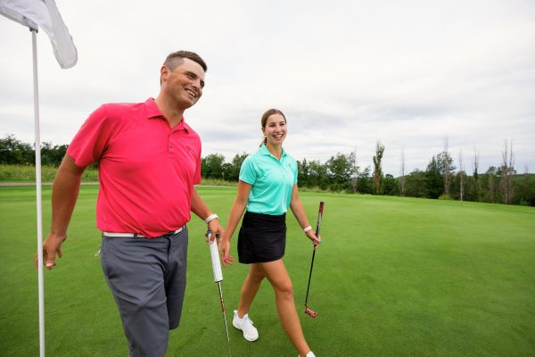 A man and a woman walk on a golf course holding golf clubs, smiling, near the hole and flag on a cloudy day.