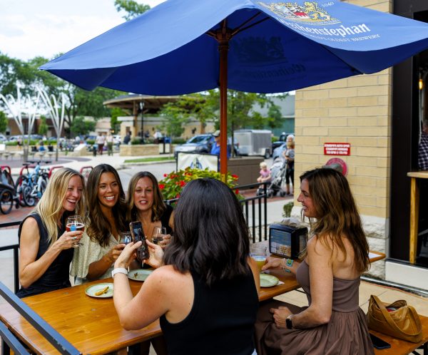 Five women sit around an outdoor table at a patio cafe, smiling and raising drinks, with food on the table and a large blue umbrella above them.