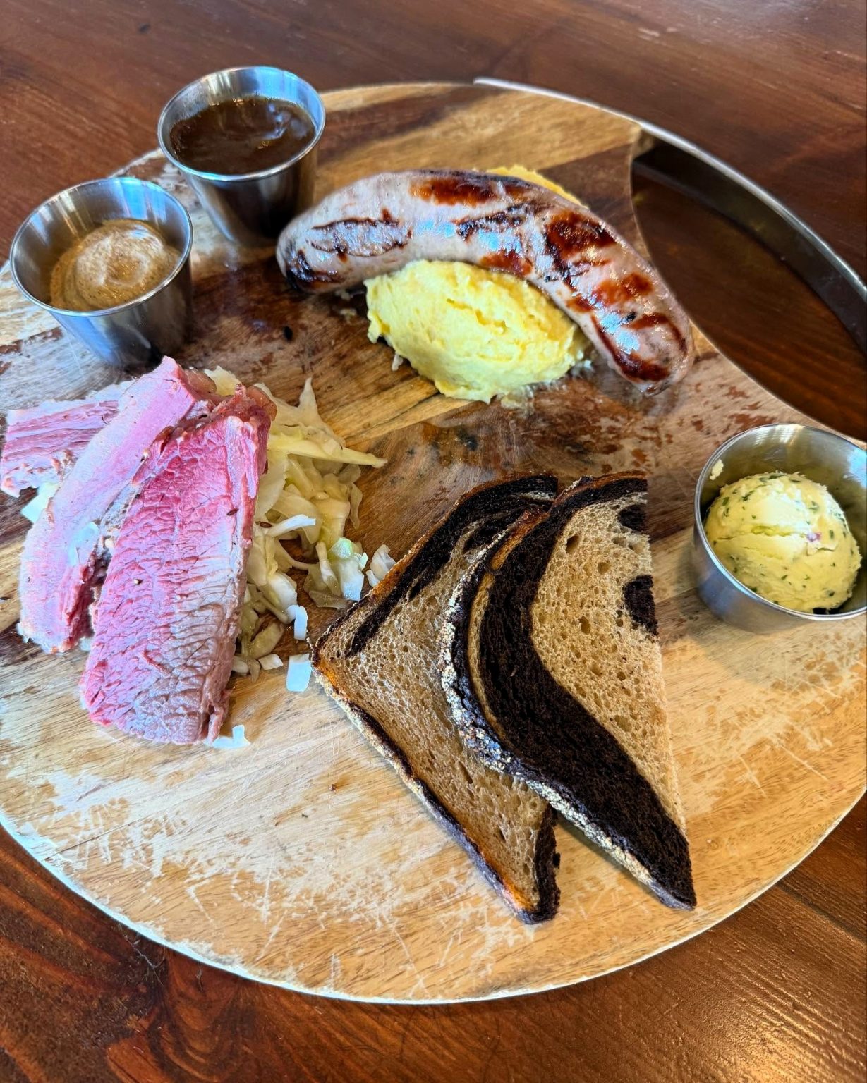 A wooden platter with grilled sausage, scrambled eggs, sliced corned beef on sauerkraut, rye bread slices, mustard, horseradish cream, and herbed butter in metal cups.