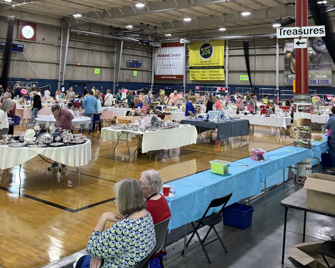 Indoor rummage sale with tables of items for sale, people browsing, and a "Treasures" sign hanging from a pole in a large gymnasium—discover hidden gems at this lively attic sale.