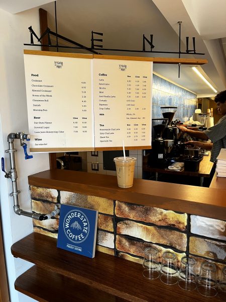 A coffee shop counter with a hanging menu, a plastic cup of iced coffee, stacked glasses, and a Wonderstate Coffee sign. A barista works behind the counter.