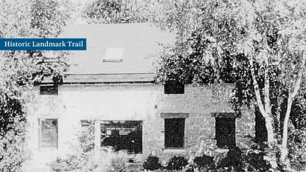 A black-and-white photo shows a brick building with three windows partially obscured by trees. A sign reads "Historic Landmark Trail" in the top left corner.