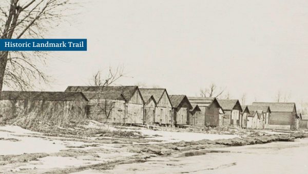 Row of small, rustic wooden cabins near a barren, snowy path and leafless trees, under a gray sky.