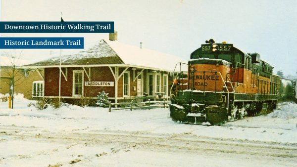 A vintage Milwaukee Road train passes a snowy Middleton depot, featuring signs for the Downtown Historic Walking Trail and Historic Landmark Trail.