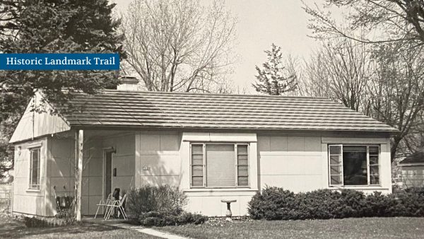 Small mid-century house with a sign reading "Historic Landmark Trail." The house has a gabled roof, two windows, a small porch with chairs, and is surrounded by trees and bushes.