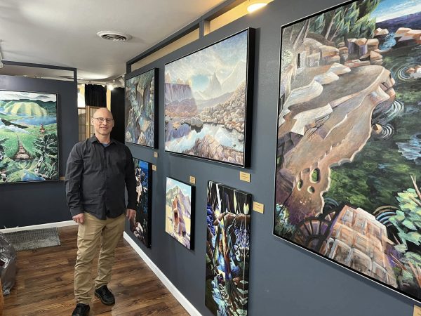 A person stands next to a blue-gray wall displaying several colorful landscape paintings in a well-lit art gallery during Gallery Night.