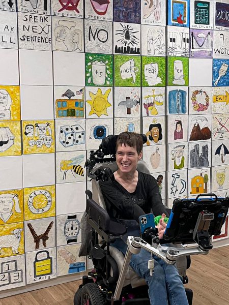 A person in a power wheelchair with assistive technology smiles in front of a colorful wall covered with hand-drawn art tiles and symbols, capturing the vibrant spirit of Gallery Night.