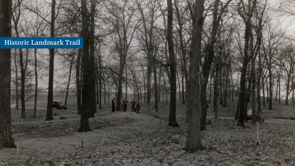 A group of people stand on a forest trail lined with bare trees. A vintage car is parked on the left. A sign reads "Historic Landmark Trail.