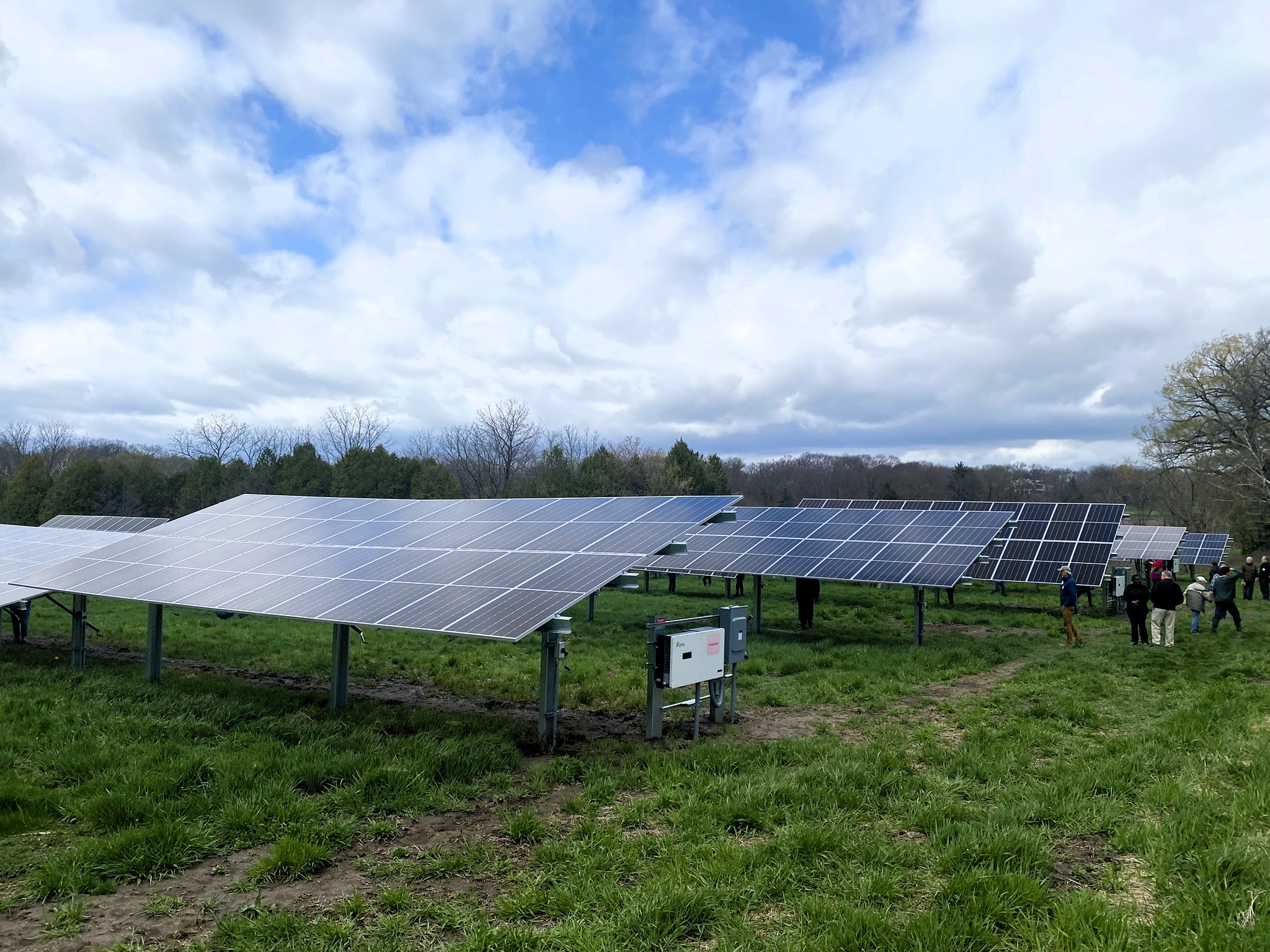 Rows of solar panels are installed in a grassy field under a partly cloudy sky; a small group of people stands nearby.