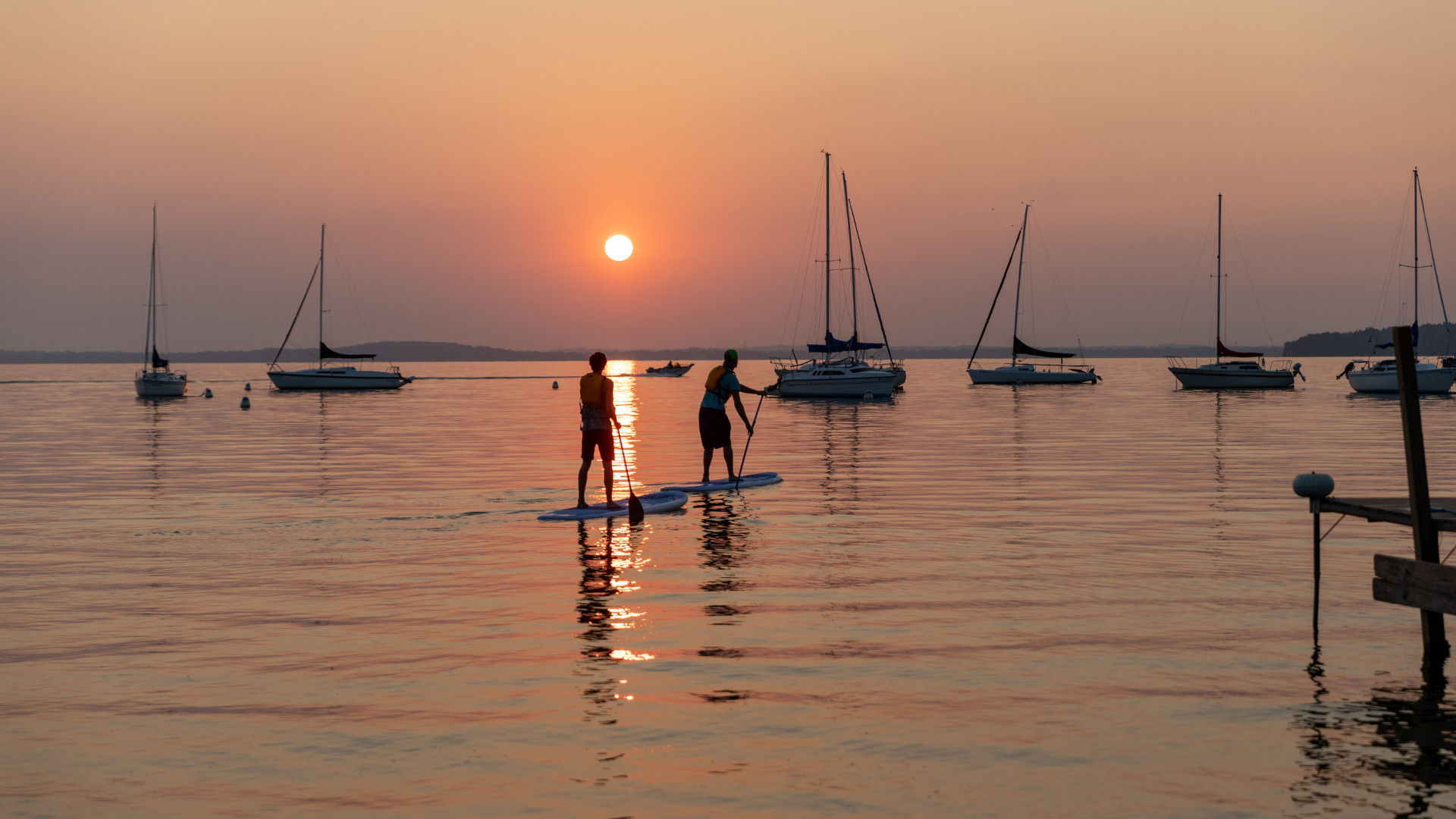 Two people paddleboarding on a calm lake at sunset, with several sailboats in the background and an orange sky.
