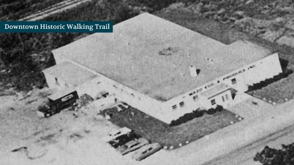 Black and white aerial view of a large, rectangular building with parked cars surrounding it. Sign reads "Downtown Historic Walking Trail.