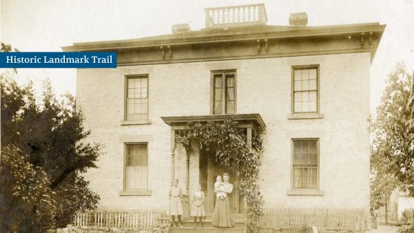 A sepia-toned photo of a two-story brick house with a central porch. Three people and a baby stand in front, with a small fence and trees nearby.