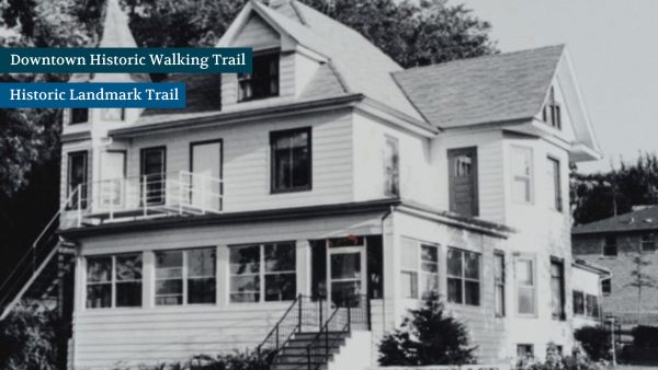 A black and white image of a two-story house with a gabled roof and a small porch, surrounded by trees. Signs indicate it's on a historic trail.