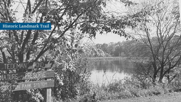 Wooden signs near trees mark the entrance to Tiedeman Pond Nature Preserve, with a small lake and more trees in the background.