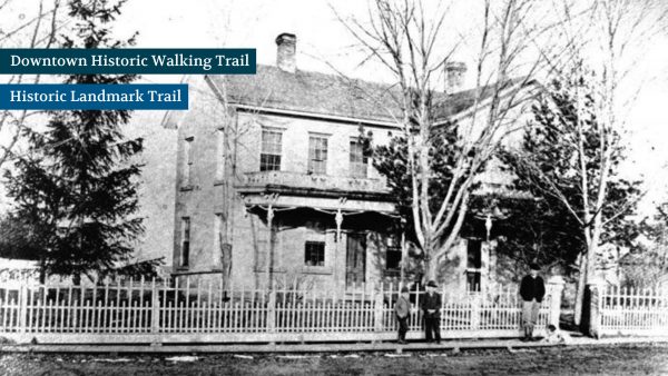 Historic black and white photo of a two-story house with a porch, surrounded by leafless trees and a picket fence. Two men and a woman stand in front of the fence.