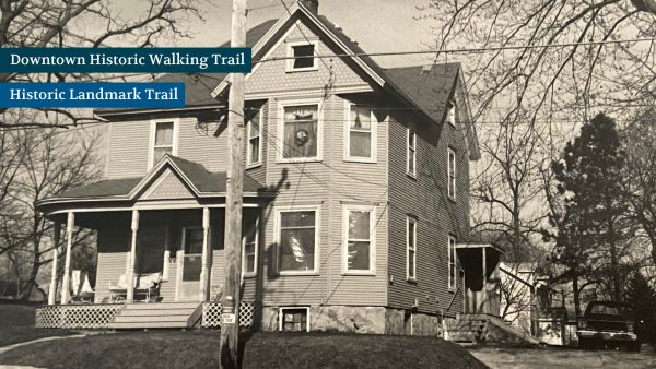 A large, two-story house with a porch and multiple windows, surrounded by leafless trees. The image is black and white.