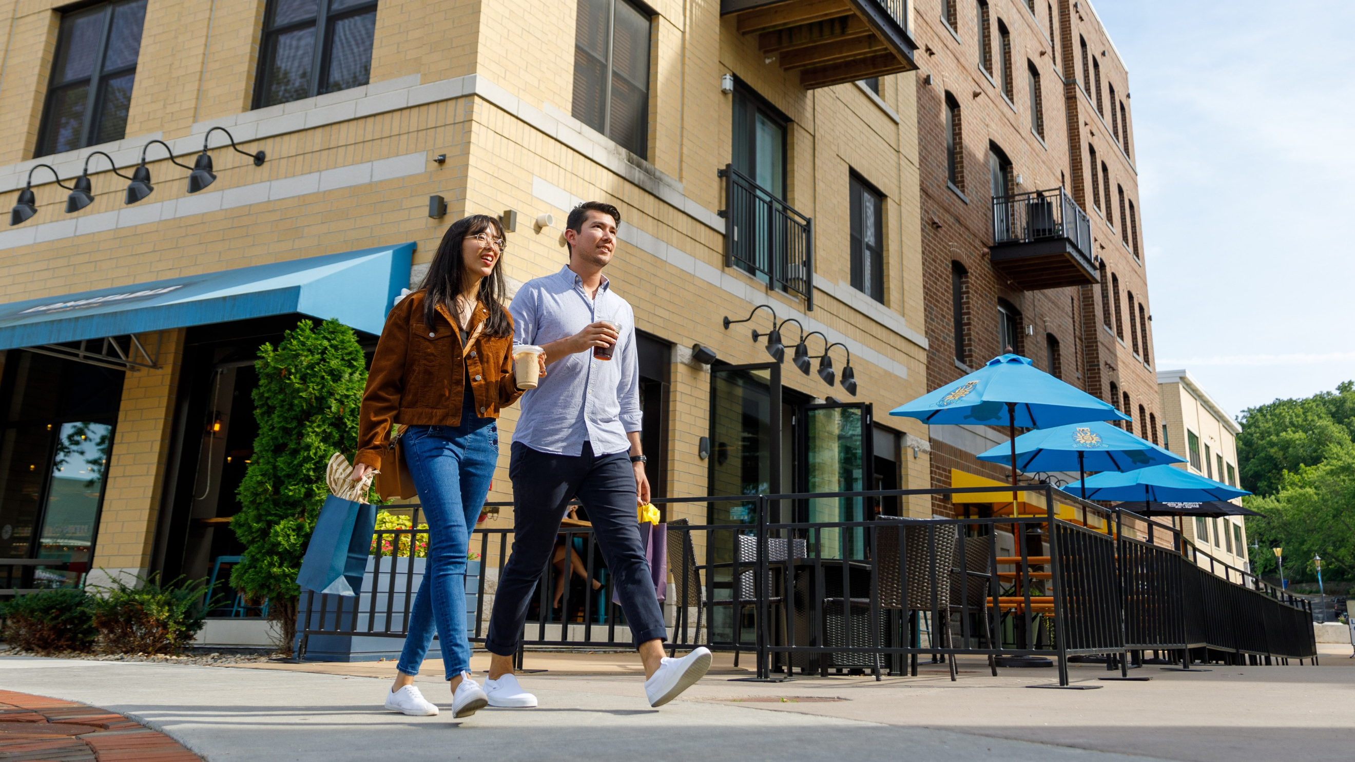 ShoppingDowntown A man and woman walk together on a sidewalk outside a building with outdoor seating and blue umbrellas on a sunny day.