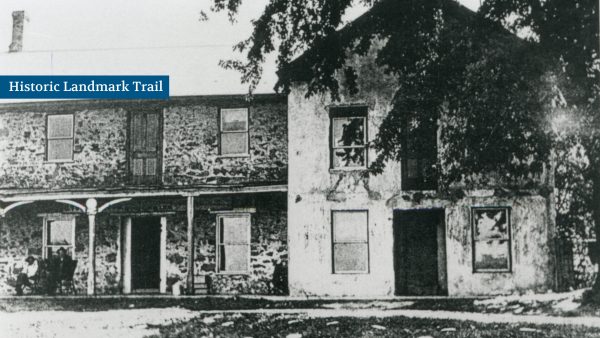 Historic stone building with two stories, a porch on the left, under a large tree. A sign reads "Historic Landmark Trail." The photo is in black and white.