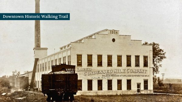 Historic building of a condensed milk company with a tall smokestack, labeled "Downtown Historic Walking Trail," and an old train car in the foreground.