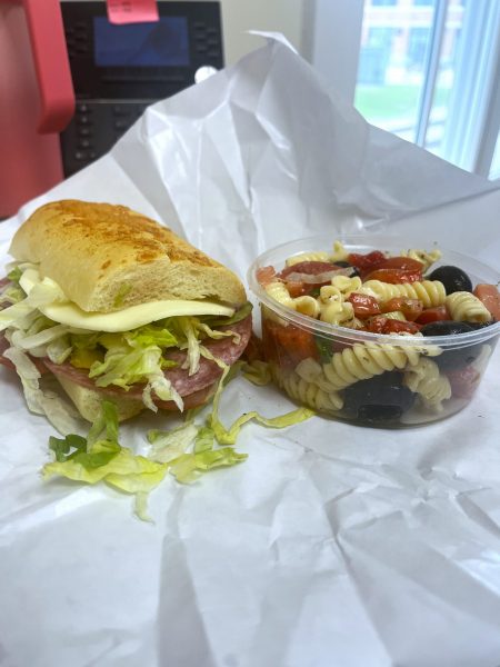A sub sandwich with lettuce, cheese, and deli meat next to a cup of pasta salad with rotini, cherry tomatoes, olives, and cheese on white paper.