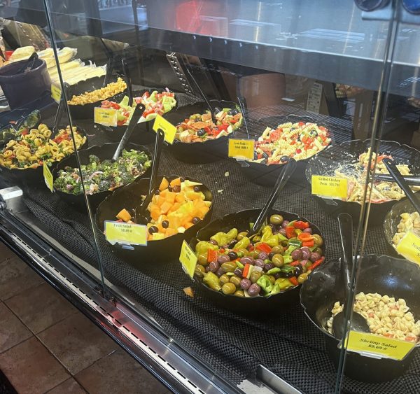 A deli salad bar display behind glass, featuring assorted salads, vegetables, pasta dishes, and cut fruit in black bowls with yellow labels.