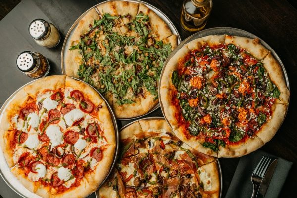 Four different pizzas topped with various vegetables, meats, and cheeses are arranged on a dark table alongside a bottle of oil and shakers of pepper and parmesan, offering flavors popular at vegetarian restaurants.