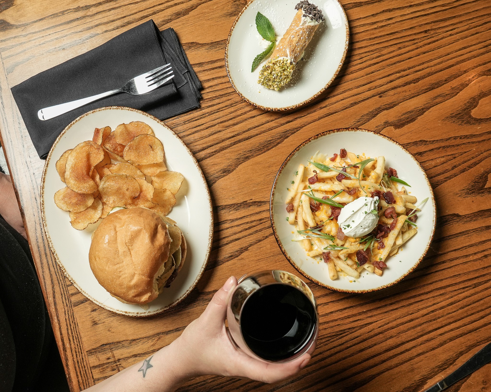 At Middleton's Restaurant Week, a person holds a glass of red wine at a table set with a burger and chips, loaded fries topped with sour cream, and a cannoli served on separate plates.
