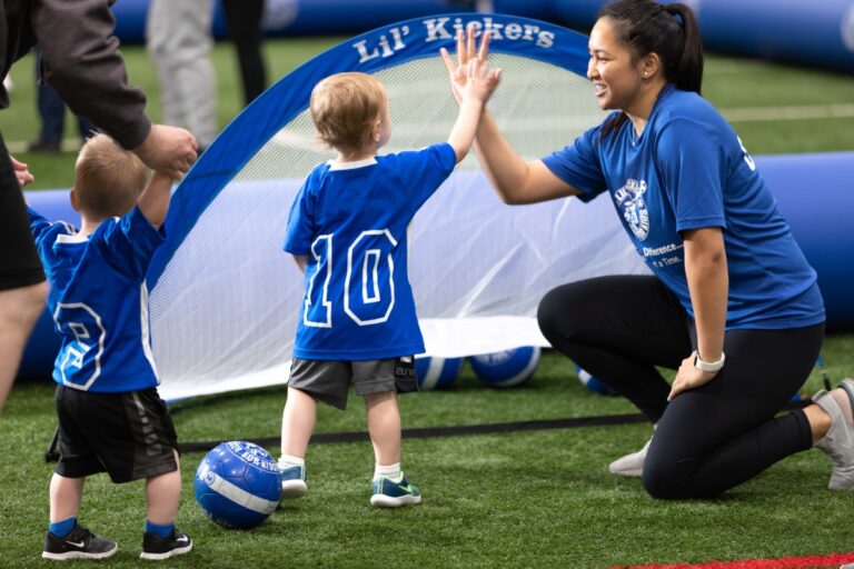 A coach kneels on artificial turf, giving a high-five to a toddler in a blue jersey while another child stands nearby, both holding soccer balls.