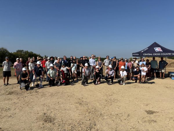 A large group of people pose together outdoors on a dirt area, some holding shovels, in front of a tent labeled "Central Cross Country Skiing" at the Middleton Nordic Center.