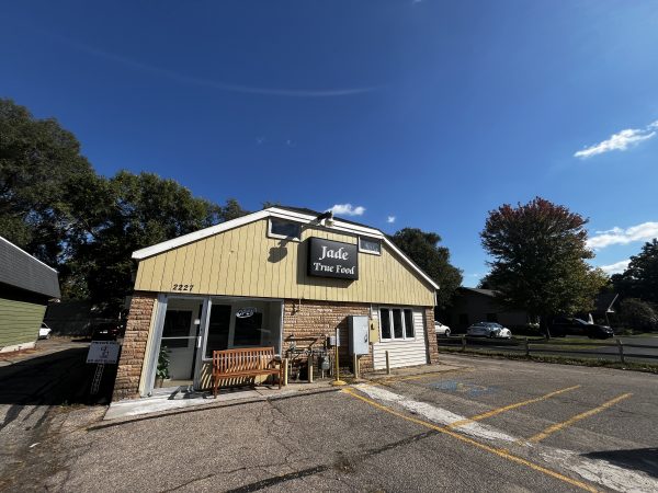 A small restaurant named "Jade True Food" with a light yellow and brick exterior, located at 2227, with outdoor seating and a parking lot in front.