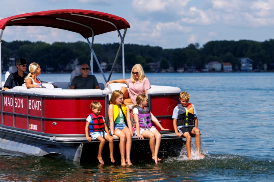 Six adults and children sit on a red pontoon boat on Lake Mendota; four children in life jackets dangle their feet in the water.