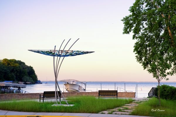 A lakeside scene with two benches facing the water, a modern metal sculpture in the foreground, a docked boat, and trees on the left and right.