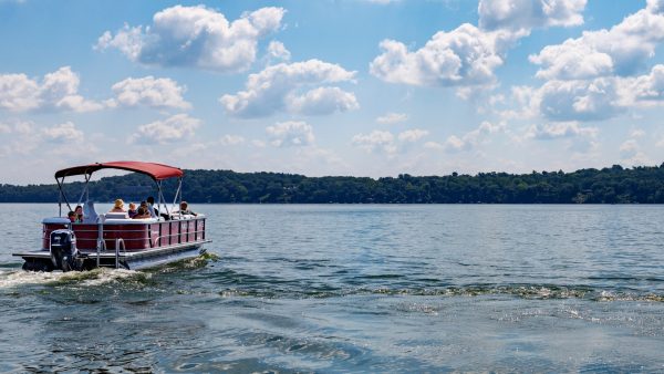 A pontoon boat with several people on board moves across a calm lake under a partly cloudy sky, with a forested shoreline in the background.
