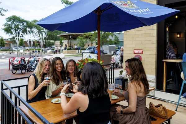 Five women sit at an outdoor restaurant table under a blue umbrella, clinking glasses and smiling, with food and drinks on the table.