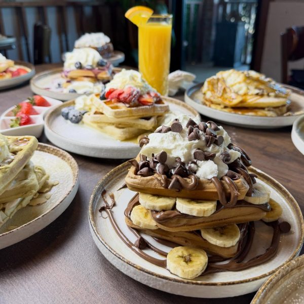 A table with plates of waffles topped with bananas, chocolate chips, strawberries, whipped cream, and a glass of orange juice in the background at La Pollera Colombiana—a delicious twist on traditional Colombian food.