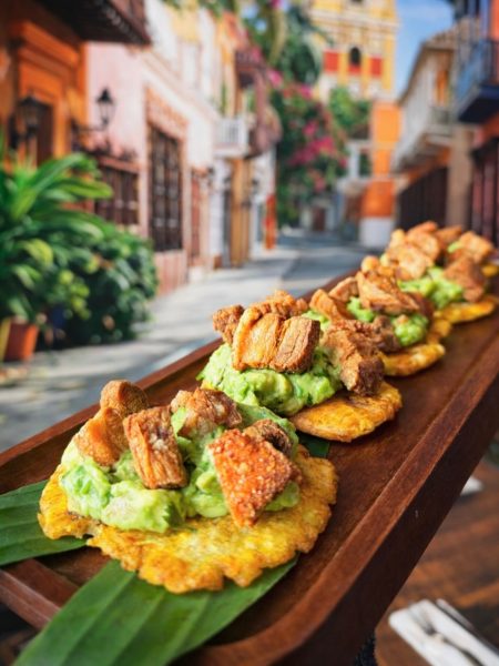 Three fried plantain tostones topped with guacamole and crispy pork pieces are arranged on a wooden tray lined with a banana leaf, capturing the vibrant flavors of Colombian food at La Pollera Colombiana, set against a colorful street scene.