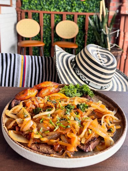 A plate of grilled steak topped with sautéed onions and herbs, served with fried plantains, captures the essence of Colombian food, sitting on a table next to a black and white striped hat and a striped cloth reminiscent of La Pollera Colombiana.