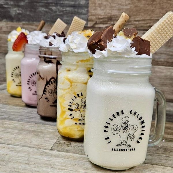 Four decorated milkshakes in mason jars with whipped cream, wafers, and toppings, lined up on a wooden surface. The jars display the "La Pollera Colombiana" logo, highlighting the creativity of Colombian food desserts.