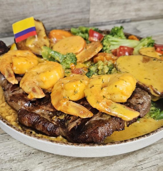 A plate from La Pollera Colombiana featuring grilled steak, shrimp in a yellow sauce, mixed vegetables, potatoes, and topped with a small Colombian flag—a true celebration of Colombian food.