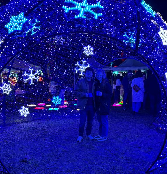 Two people stand inside a dome of blue holiday lights and snowflake decorations at night, with other people, illuminated displays, and nearby vegetarian restaurants in the background.