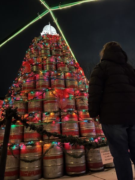 A person stands in front of a large Christmas tree structure made of beer kegs, decorated with colorful lights and red bows, at night, near a bustling area filled with vegetarian restaurants.