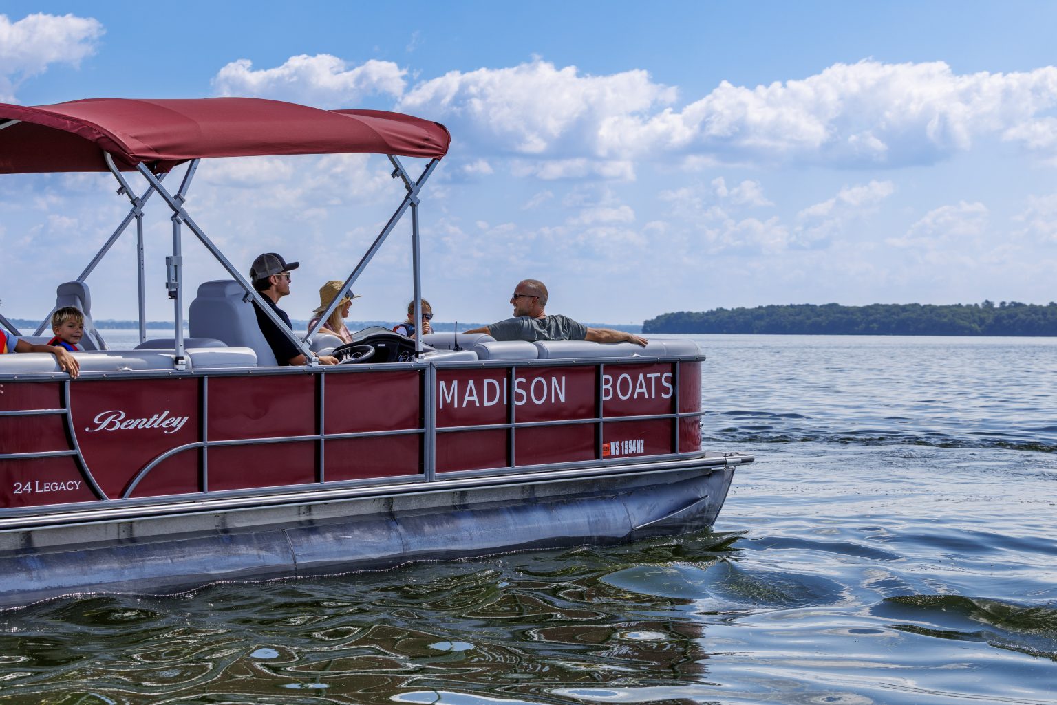 A red Madison Boats pontoon with four people on board moves across a calm lake under a partly cloudy sky.