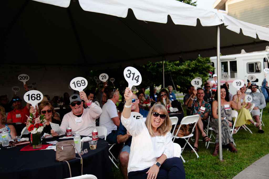 A group of people sit under a tent, raising numbered paddles at what appears to be an outdoor auction event. A smiling woman in front holds up paddle 314.