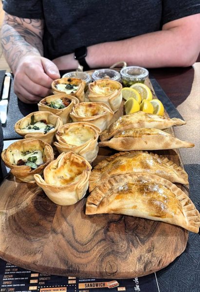 A wooden board with baked empanadas, mini quiches, lemon wedges, and small sauce containers sits in front of a person with tattooed arms at Toro Y Pampa, an Argentinian restaurant.