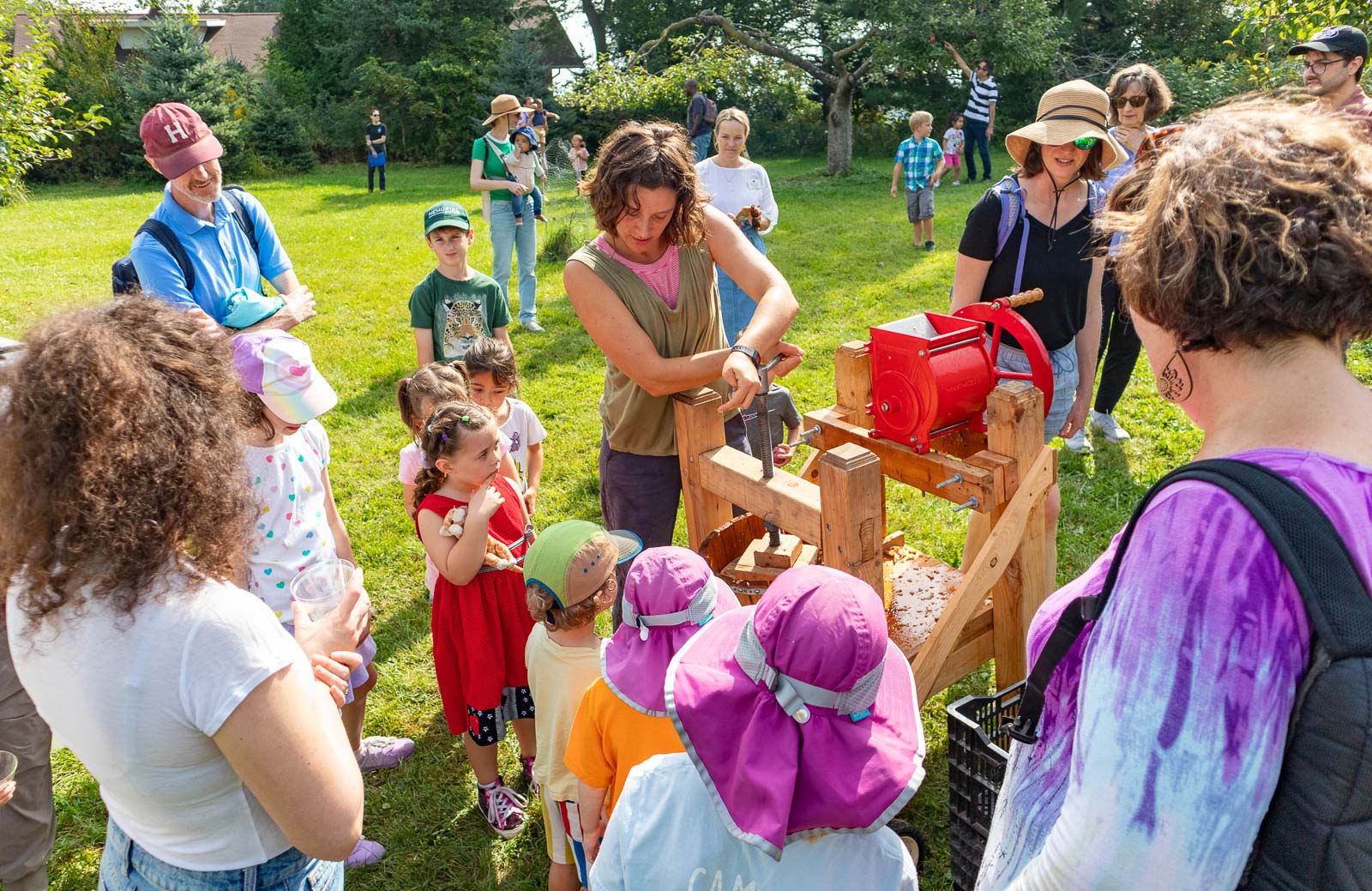 A group of children and adults watch a woman operate a hand-cranked apple press outdoors on a sunny day.