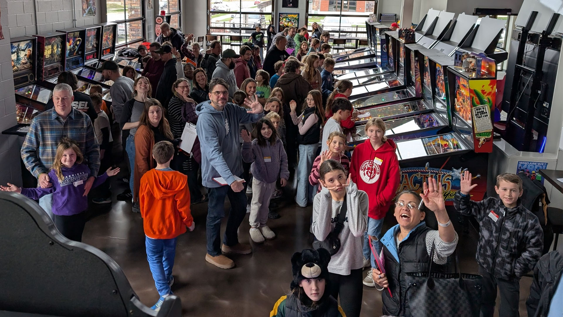 A large group of people, including children and adults, gather inside an arcade filled with pinball machines, many smiling and waving at the camera.