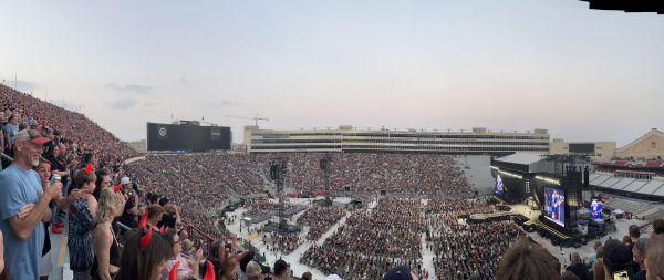 A large crowd fills a stadium for a concert, reminiscent of the exciting atmosphere found at Kohl Center concerts. The stage, screens, and lighting are set up, with the audience seated and standing across the venue. The sky is clear in the background.